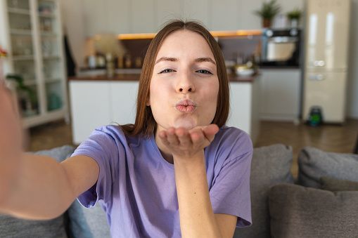 Attractive young woman using camera of mobile phone taking selfie at home blows a kiss to boyfriend or friends