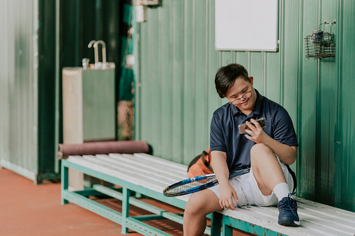 young asian Chinese down syndrome male tennis player resting at bench while using handphone