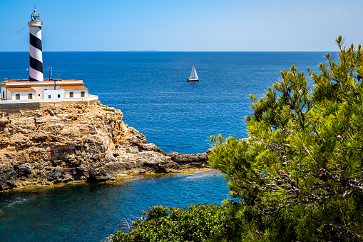 Idyllic Mallorca bay Es Mular with the lighthouse Faro de Cala Figuera in front of a sailboat on the mediterranean sea and islands of Cabrera national park on the horizon with pine tree in foreground.