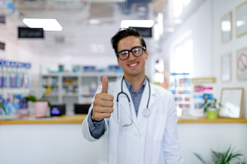 A young Caucasian male doctor is standing in a pharmacy and looking at the camera while cheerfully giving thumbs-up.