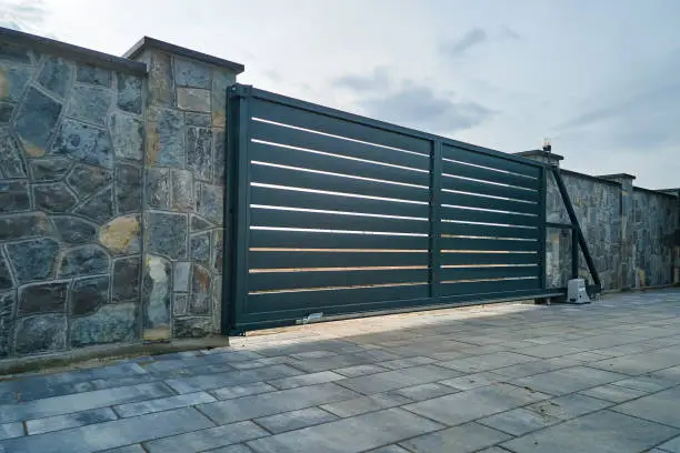 Photo of Wide automatic sliding gate with remote control installed in high stone fense wall. Security and protection concept