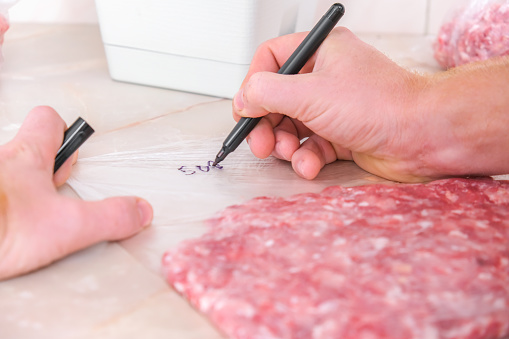 A man signs with a marker the date of production and a gram of minced pork and beef laid out in bags for storage in the freezer. Product labeling.