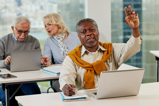 Las personas mayores se sientan en un aula y hablan sobre el proyecto en la computadora portátil mientras un estudiante multicultural de último año en primer plano hace preguntas. photo