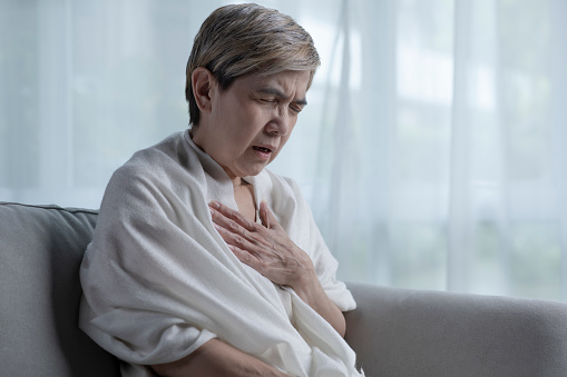 Senior Asian woman suffering from heart attack while alone at home. Woman having chest pain from cardiac arrest. Middle-aged woman putting hands on chest having trouble breathing from heart condition. Mature woman suffering from stroke or cardiovascular disease in health care wellness and medical concept.