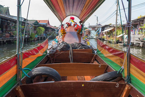 Traditional wooden long tail boat floating on canal water flowing between small shabby residential houses in Thonburi area of Bangkok