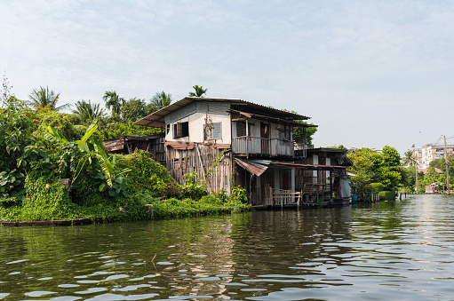 Old shabby residential house built on khlong flowing amidst lush tropical plants against cloudy blue sky in Thonburi district of Bangkok