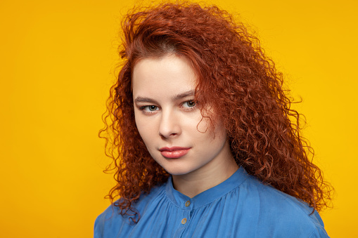 Young woman with long red hair in blue dress close-up studio portrait on yellow background