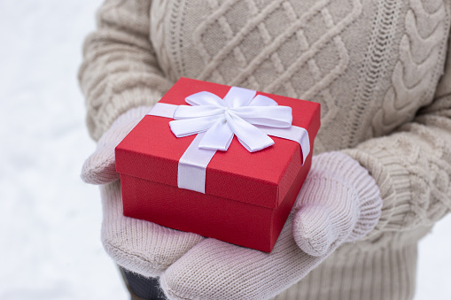 A girl in a white sweater holds a red gift box in white mittens in her hands. Close up. No face