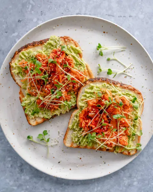 Overhead view of a plate with two toasts with mashed avocado, kimchi, hemp seeds and microgreens, one toast partly eaten