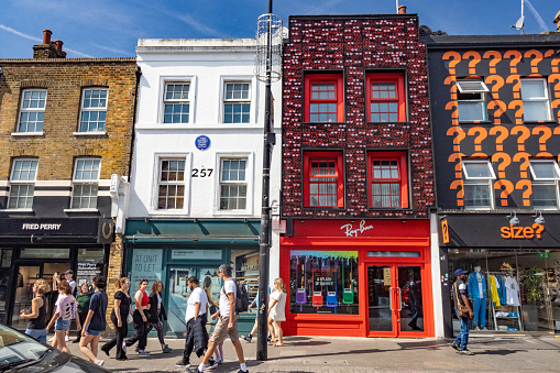 People walking past the Ray-Ban Store on Camden High Street in Borough of Camden, London. Other stores are visible. The blue plaque honours pugilist Tom Sayers (1826-1865), a boxing legend of the Victorian era who died at 39.