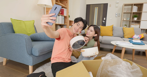 happy asian couple unpack parcel from online shopping delivery service holding new pot and use phone take picture in living room at home