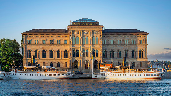 Nationalmuseum, or National Museum of Fine Arts, located on the peninsula Blasieholmen in central Stockholm, Sweden - View from Stockholms stromo with ferries parked in front at sunset in a summer day