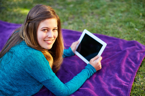 Happy, relaxed young woman with long hair using digital tablet outdoors amid greenery as she lies on her front on a blanket on a lawn.