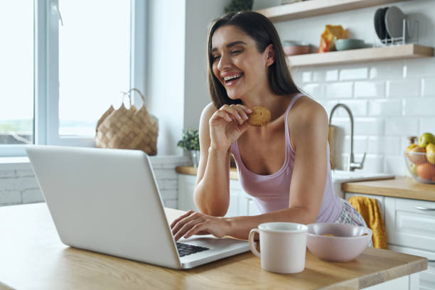 istock Happy young woman using laptop and eating cookies while sitting at the kitchen counter 1411973549