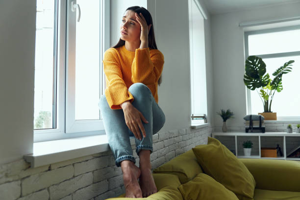 istock Depressed woman touching head and looking through a window while sitting on the window sill 1411973846