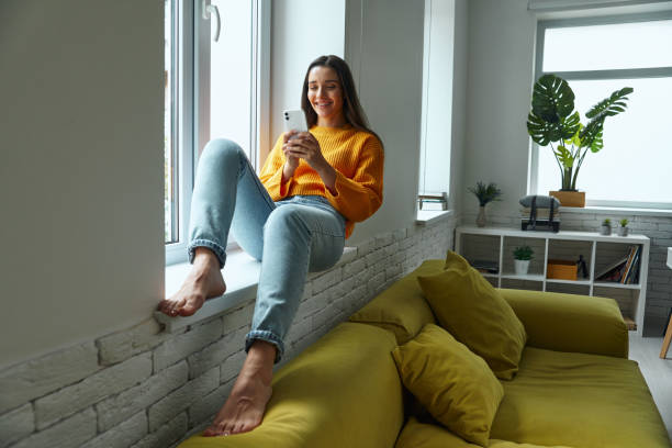 istock Happy young woman using smart phone while sitting on the window sill at home 1411973945