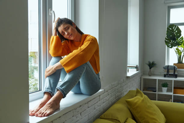istock Frustrated young woman leaning head on hand while sitting on the window sill 1411974498