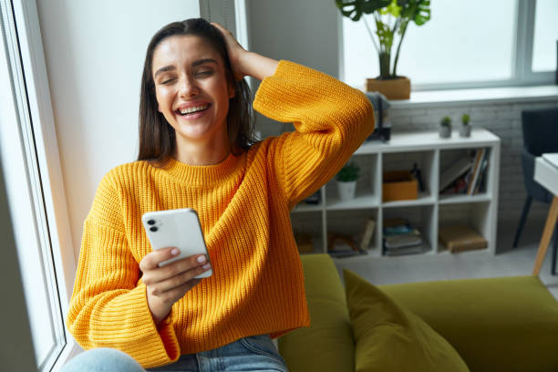 istock Beautiful young woman using smart phone and smiling while sitting on the window sill 1411976054