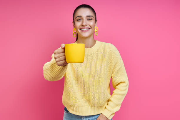 Attractive young woman in yellow sweater holding coffee cup and smiling against colored background stock photo