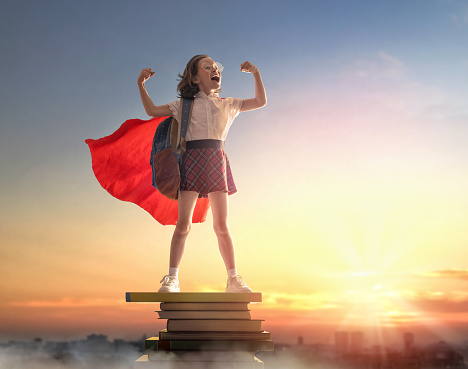 Back to school! Happy cute industrious child standing on the tower of books on background of sunset sky. Girl is wearing superhero cape. Concept of success education and reading. The development of the imagination.