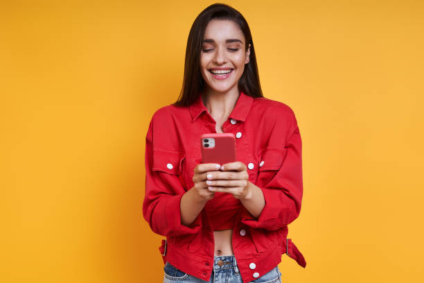 istock Happy young woman using smart phone while standing against yellow background 1411976472