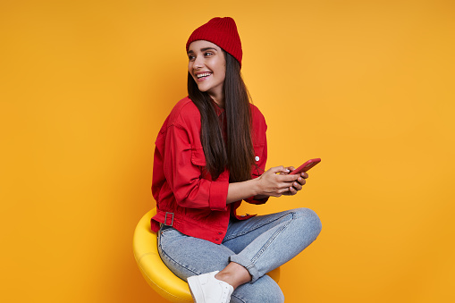 Happy young woman using smart phone while sitting on the chair against yellow background