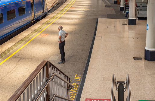York, UK. July 27, 2022.  A man stands on a railway station platform watching a train speed by. Handrails are in the foreground.