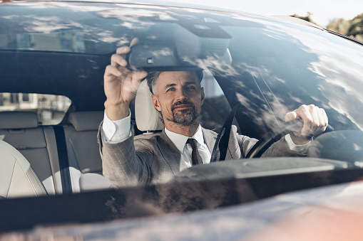 Confident man in formalwear adjusting rear view mirror while sitting on the front seat of a car