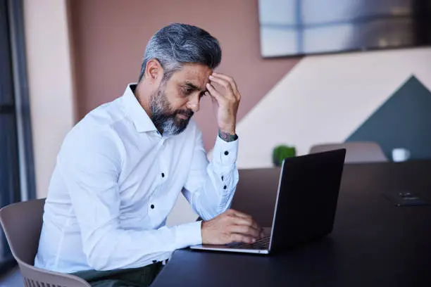 Businessman looking stressed out while working on a laptop at a table in the boardroom of an office