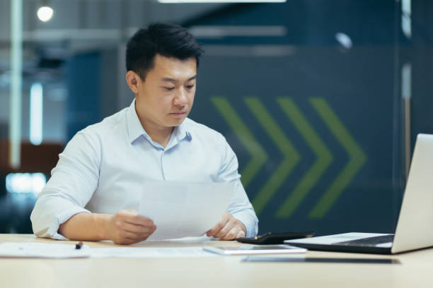 istock Young Asian male working with documents and calculator. Sitting with a laptop in a modern office 1411984143