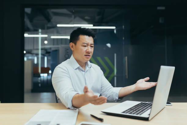 un homme d’affaires asiatique confus, assis devant un ordinateur portable dans le bureau, parle sur un appel vidéo, écarte les mains - laptop japanese ethnicity businessman desk photos et images de collection