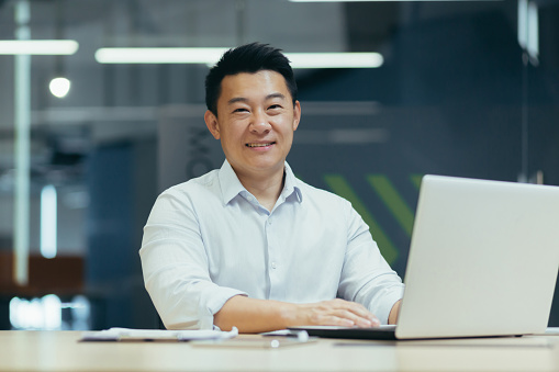 Portrait of a young Asian man, looking at the camera, smiling. Sitting at the desk in the office, working, studying with a laptop online