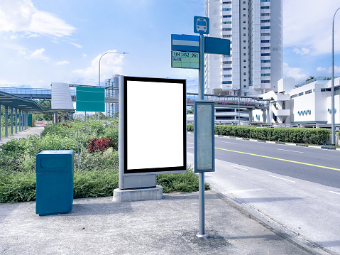 Blank vertical advertising poster banner mockup at empty bus stop shelter by main road, greenery behind; out-of-home OOH vertical billboard media display space