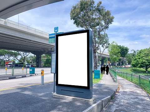Blank vertical advertising poster banner mockup at empty bus stop shelter by main road, greenery behind. Out-of-home OOH vertical billboard media display space under expressway highway