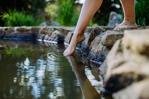 An unrecognizable young woman is dipping her foot in cool water of pond, refreshing and hardening concept.