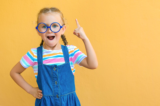Portrait of child girl in glasses with finger pointed up. Little kid in pose has idea isolated on yellow blackboard copy space. Back to school.