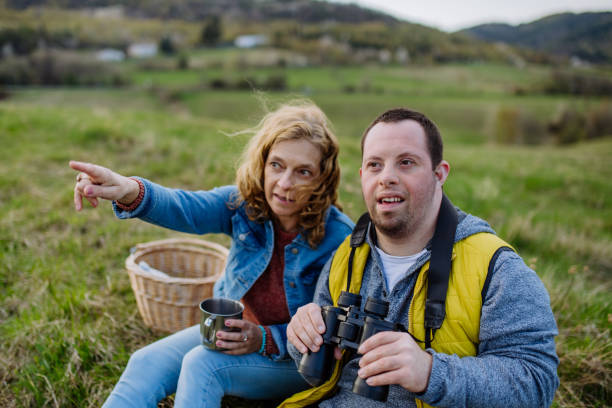 Portrait of happy young man with Down syndrome with his mother resting in nature, sitting and looking at view with binoculars. A portrait of happy young man with Down syndrome with his mother resting in nature, sitting and looking at view with binoculars. disabled person hiking outdoors stock pictures, royalty-free photos & images