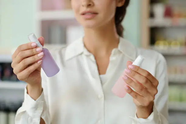 Hands of young shopper holding two plastic bottles with liquid nailcare products in cosmetic shop while deciding which one to choose