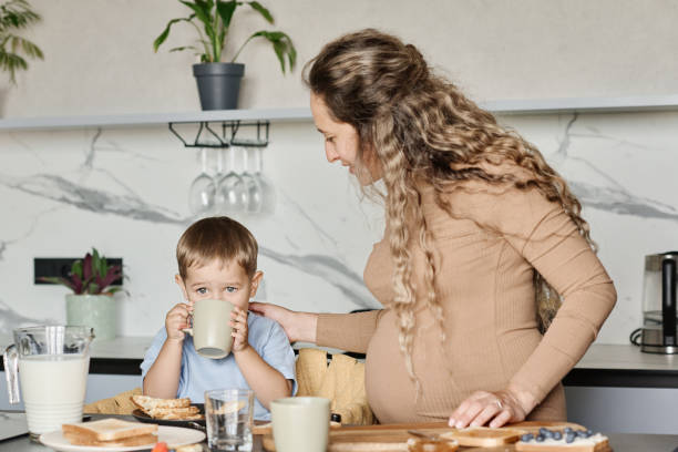 Young pregnant woman standing next to her little son drinking milk or tea Young pregnant woman standing next to her little son with mug drinking milk or tea while having homemade sandwiches for breakfast morning time management for preschool for babies stock pictures, royalty-free photos & images