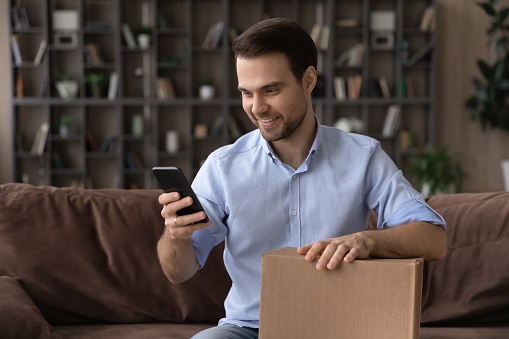 Smiling man using smartphone, holding cardboard box, unpacking parcel browsing mobile device apps, blogger shooting unboxing video, happy customer satisfied by good delivery service, sitting on couch