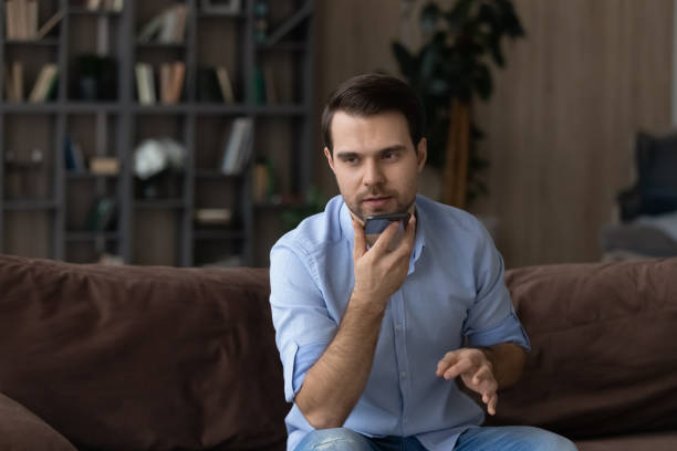 istock Serious man recording audio message on smartphone, sitting on couch 1412021239