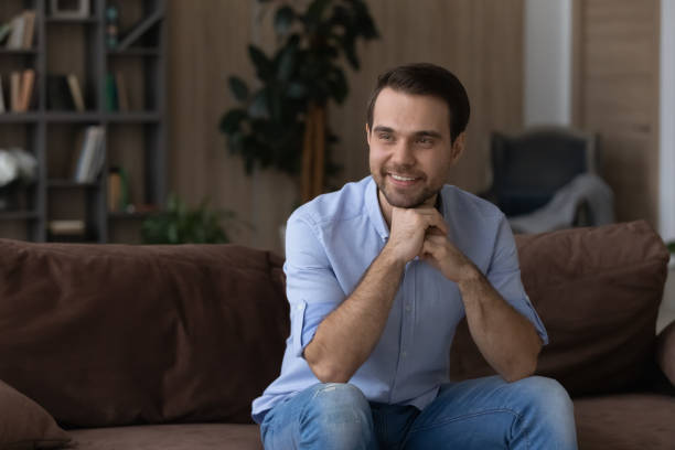 istock Smiling dreamy man sitting on couch, looking in distance 1412021260