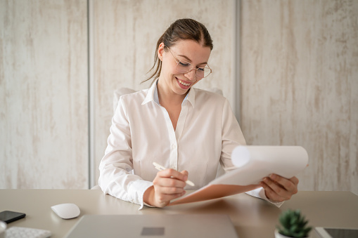 One woman female caucasian entrepreneur businesswoman or secretary sitting at her office at desk work happy smile checking paper documents contract wear white shirt copy space