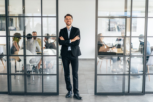 Shot of a businessman standing in the modern office with his arms crossed looking confident and smiling at the camera, colleagues behind working