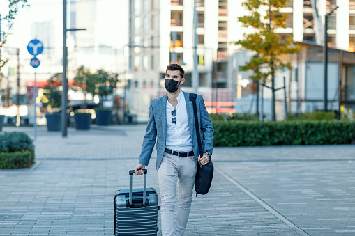 A young Caucasian businessman wearing a face mask is walking down the street and carrying a suitcase on his business trip.