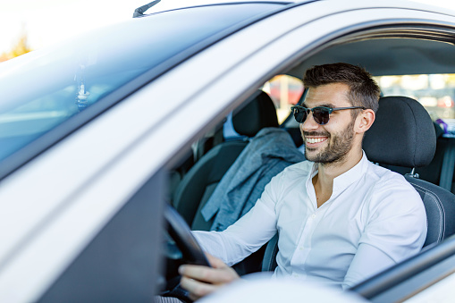 A young Caucasian man wearing sunglasses is enjoying a morning car ride to work with a big smile on his face.