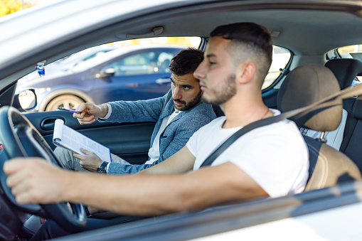 A young Caucasian male driving instructor is sitting in a car and giving advice to a young Caucasian man taking the driver's licence test.