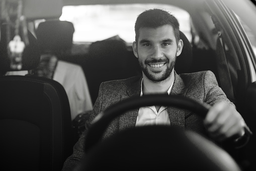 A young Caucasian businessman is sitting in his car with his hand on the wheel, and looking at the camera with a warm smile on his face.