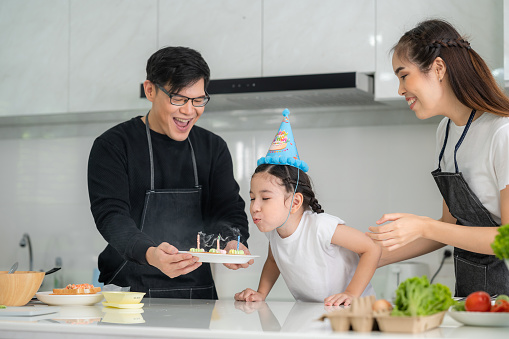 Little Asian girl celebrating her birthday with parents blowing out the candles on her cake in kitchen at home. Happy family time