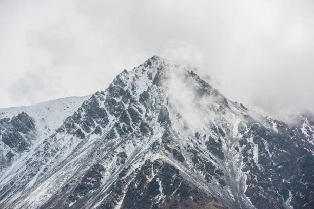 istock Awesome landscape with high snowy mountain peaked top with sharp rocks in low clouds. Dramatic view to snow mountain pointed peak in low cloudy sky. Monochrome scenery with white snow on black rocks. 1412087939